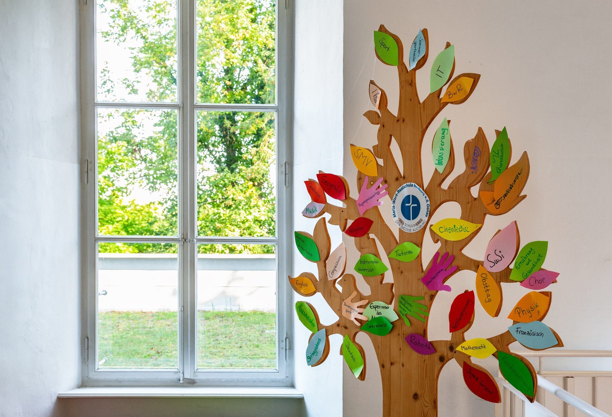Holzbaum mit Fächern und Angeboten der Maria-Ward-Realschule Neuburg im Treppenhaus
